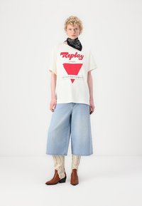 T-shirt w kolorze off-white z czerwoną grafiką, połączony z jasnoniebieskimi, szerokimi szortami dżinsowymi. Brązowe botki za kostkę i wzorzyste skarpetki do kolan dopełniają wygląd.