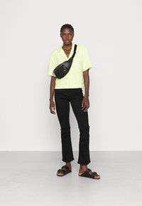 Camisa de manga curta, verde limão, jeans flare pretos, bolsa de tiracolo preta e sandálias pretas com sola de madeira. Fundo branco liso.