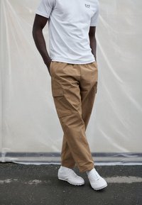 Bež cargo hlače sa bočnim džepovima, lagana tkanina, ravni kroj, u kombinaciji s bijelom majicom i bijelim sportskim tenisicama.