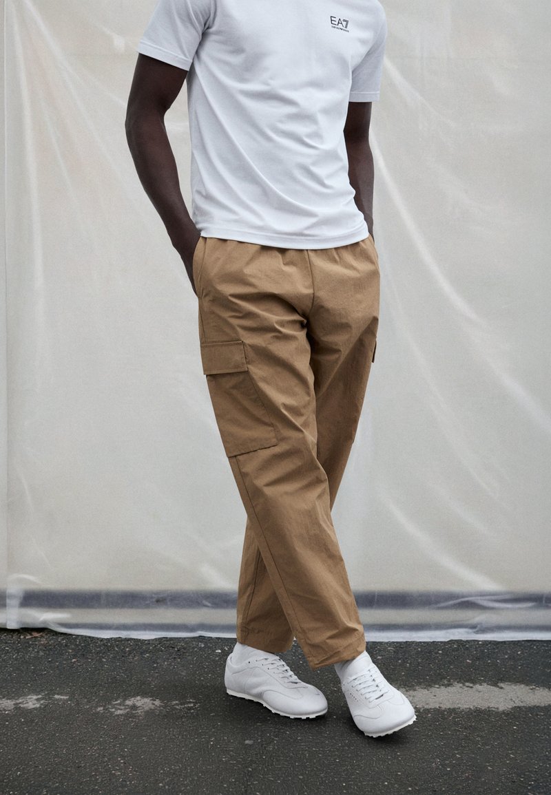 Bež cargo hlače sa bočnim džepovima, lagana tkanina, ravni kroj, u kombinaciji s bijelom majicom i bijelim sportskim tenisicama.
