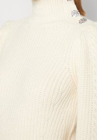 Maglione crema a collo alto con texture a coste, dotato di dettagli decorativi in argento sulla spalla. Tessuto morbido, design aderente.
