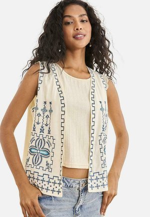 Mujer con cabello largo y rizado, que lleva un chaleco sin mangas color crema con bordado azul sobre una camiseta sin mangas beige y unos vaqueros azul claro.