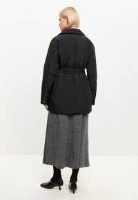 Chaqueta de plumas negra con cintura ceñida por un cinturón, gran cuello y puños abotonados. Llevada sobre una falda larga gris y medias negras.