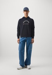 Felpa con cappuccio blu navy con testo bianco e accenti rossi, abbinata a jeans a gamba larga blu con tasche cargo laterali e sneakers bianche.