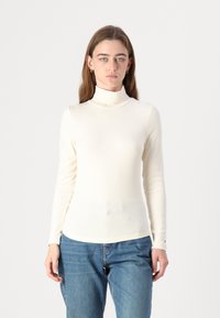 Kremfarget høyhalset skjorte med ribbestruktur, lange ermer og tettsittende design; kombinert med løse blå denimjeans.