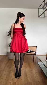Γυναίκα με κόκκινο σατέν φόρεμα και μαύρο καλσόν που στέκεται σε εσωτερικό χώρο σε ξύλινο πάτωμα, με μια ψάθινη καρέκλα και μια γλάστρα στο παρασκήνιο.