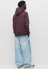 Felpa con cappuccio color marrone scuro con polsini a coste, abbinata a pantaloni cargo larghi blu chiaro con grandi tasche. Indossata con sneakers a fantasia.