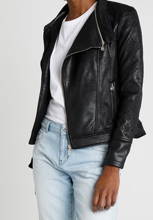 Personne portant une veste en cuir noire texturée sur un t-shirt blanc et un jean bleu clair, debout devant un fond blanc.