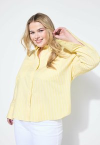 Gestreiftes Hemd in Gelb und Weiß mit Kragen, langen Ärmeln und vorderen Taschen, kombiniert mit weißen Hosen. Glatte Stoffstruktur.