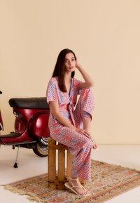 Une combinaison à imprimé floral rouge et bleu, avec des manches courtes, une taille à nouer et des jambes larges. Modèle assis sur une caisse en bois à côté d'un scooter rouge.