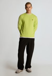 Felpa a maniche lunghe verde lime con colletto bianco e logo giallo, abbinata a pantaloni neri larghi e scarpe grigie.