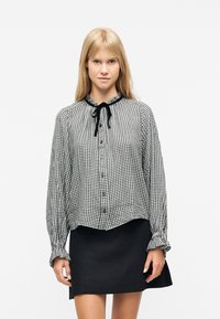 Femme blonde portant un chemisier à carreaux noir et blanc avec un nœud noir au col et une jupe noire courte sur un fond blanc.