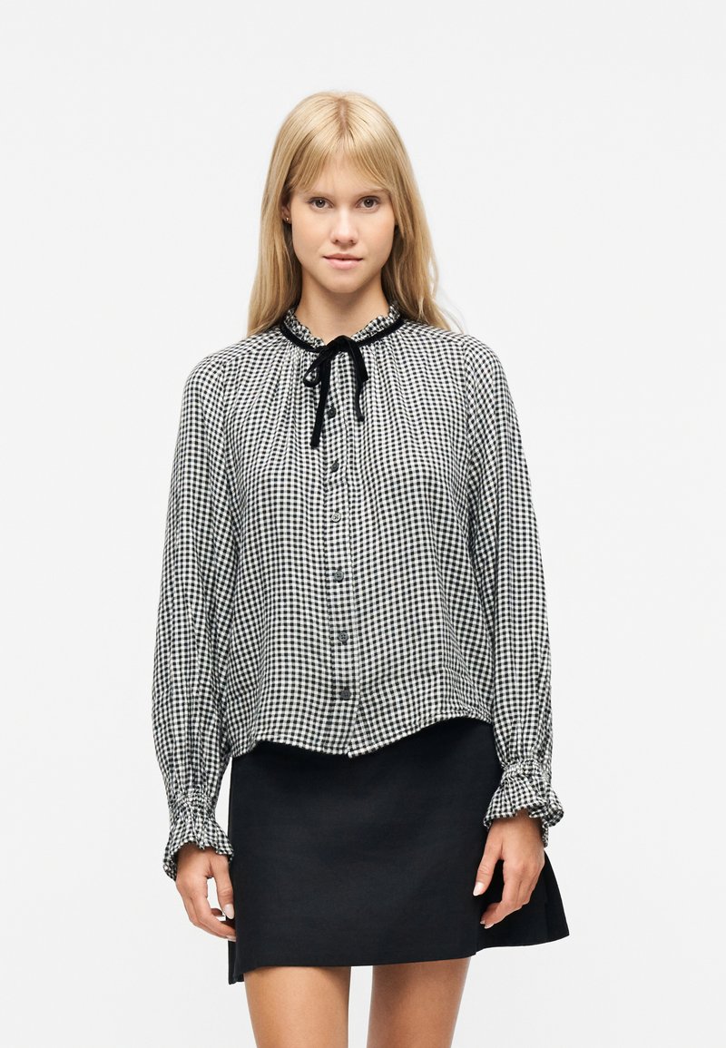 Femme blonde portant un chemisier à carreaux noir et blanc avec un nœud noir au col et une jupe noire courte sur un fond blanc.