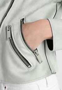 Ljusgrön faux suede jacka med silverdragkedjor, som har två sidofickor med dragkedja och en elegant design. Närbild på fickdetalj synlig.