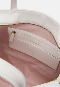 Interno di una borsa bianca con rivestimento rosa chiaro, dotato di una tasca con zip e scomparti per carte. Logo del marchio in oro visibile. Texture liscia.