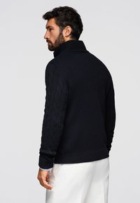Homme barbu portant un pull col roulé en tricot noir et un pantalon blanc, se tenant de côté contre un fond uni.