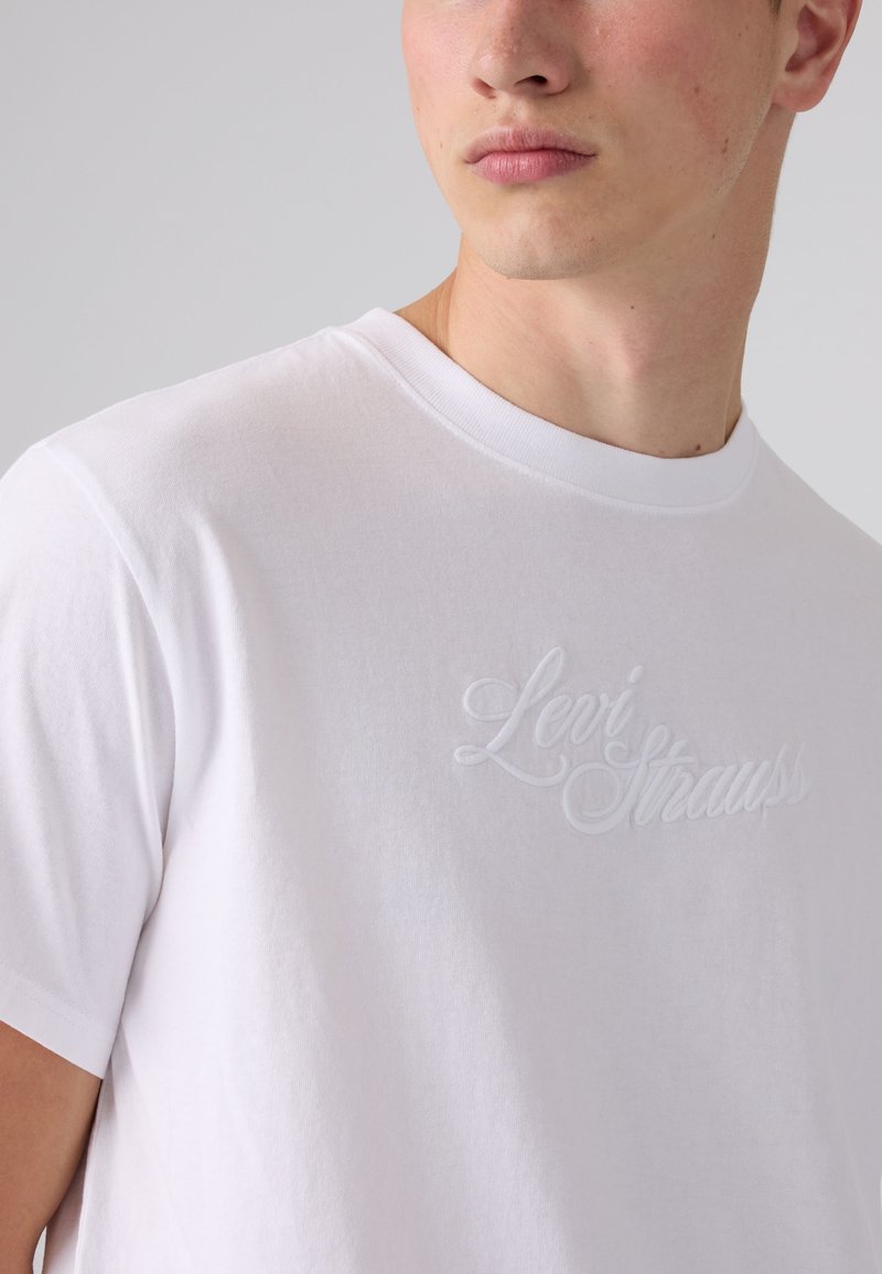 Man wearing white crew neck t-shirt with embossed Levi Strauss logo on chest against plain background.