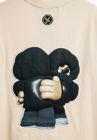 Tela beige con un gran gráfico bordado negro de dos figuras estilizadas abrazándose, con detalles texturizados y un emblema negro redondo encima.