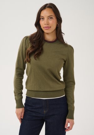Joven mujer con cabello largo y ondulado, vestida con un suéter verde oliva y vaqueros azul oscuro, de pie frente a un fondo claro y liso.