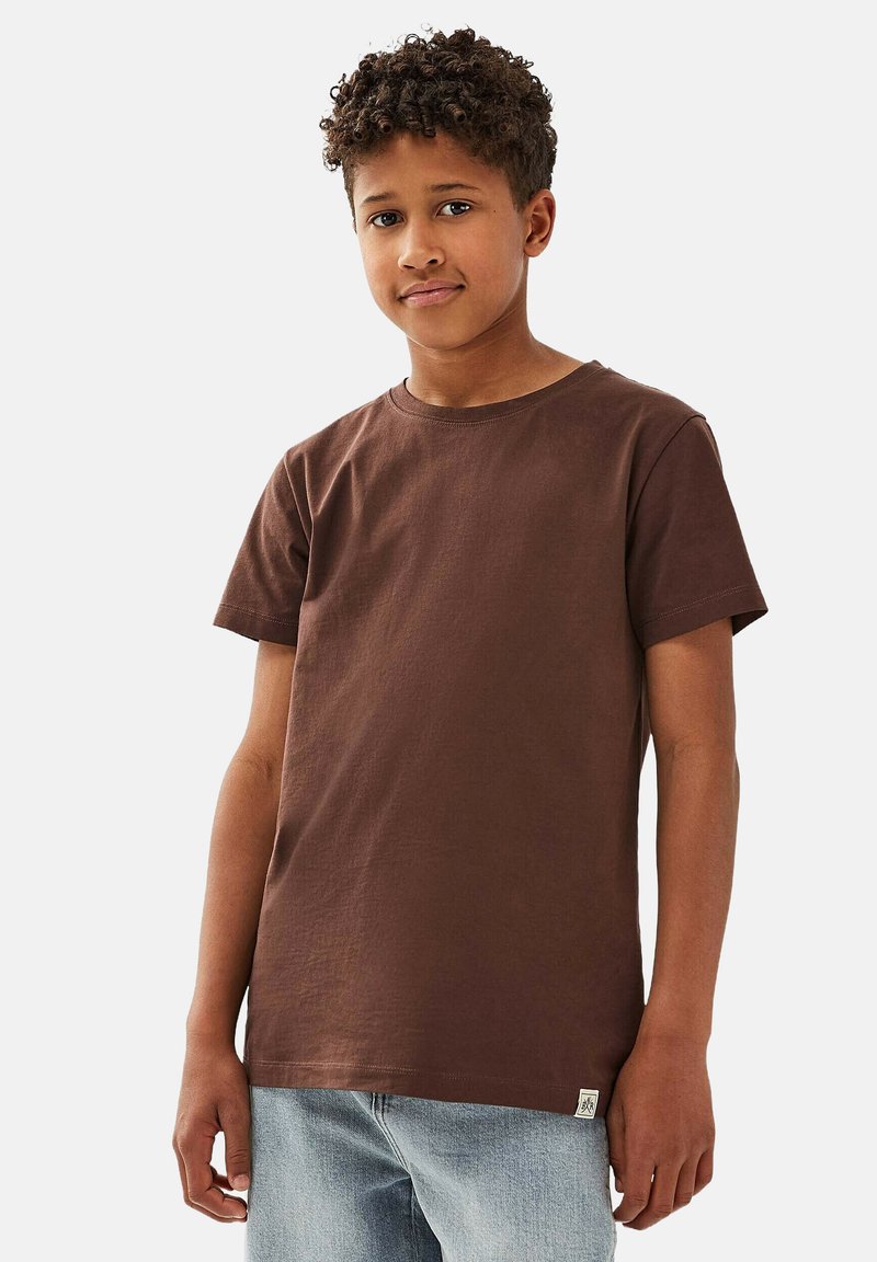 Poika, jolla on kiharat hiukset, pukeutuneena yksinkertaiseen ruskeaan lyhythihaiseen t-paitaan ja vaaleansinisiin farkkuihin, seisoo valkoista taustaa vasten.