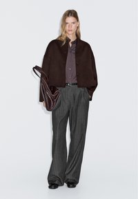 Donna con giacca buttonata marrone scuro, camicia viola, pantaloni a gamba larga grigio rigato, scarpe nere, che porta una grande borsa di pelle marrone.