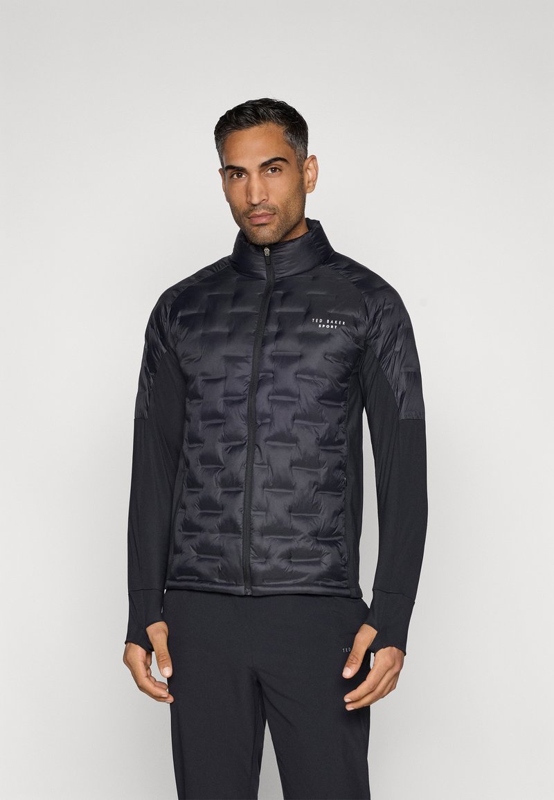 Μαύρο τζάκετ puffer με ραφές και ψηλό γιακά, διαθέτει κλείσιμο με φερμουάρ μπροστά και λογότυπο στη θήκη. Μακριά μανίκια από αντίθετο ύφασμα.