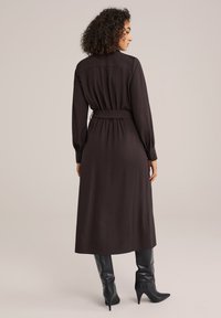 Robe longue à manches longues marron foncé avec une taille ceinturée, présentant une texture lisse et un ourlet midi. Accompagnée de bottes noires jusqu'aux genoux.