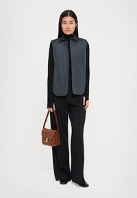 Grå ermeløs vest med krage-design over en svart turtleneck, parret med vide svarte bukser og en brun lær skulderveske.