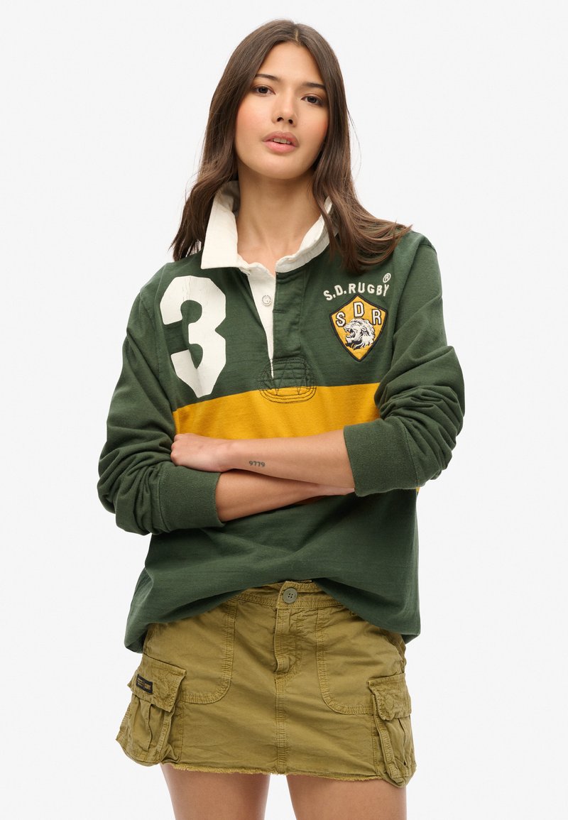 Grünes Rugbyshirt mit einem weißen Kragen, ausgestattet mit einem gelben Streifen und einer großen "3" am linken Ärmel. Kombiniert mit einem khakifarbenen Cargo-Rock.