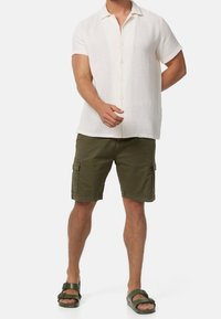 Hombre con camisa blanca de manga corta abotonada, pantalones cortos cargo verde oliva y sandalias oscuras de doble tira, de pie contra un fondo blanco.