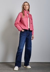 Veste rose cropped avec un col haut et des boutons pression, associée à un jean évasé bleu foncé et des baskets blanches sur un fond gris.