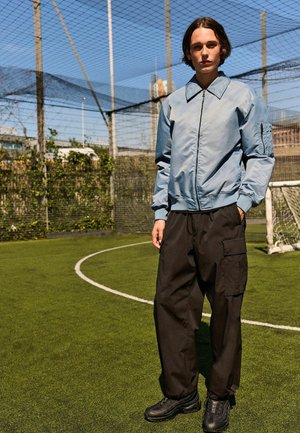 Helleblaue Bomberjacke mit Reißverschluss vorne, gerippten Bündchen und einer aufgesetzten Tasche. Kombiniert mit schwarzen Cargo-Hosen und schwarzen Sneakers auf einem grünen Feld.