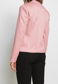 Rosa strukturerad blazer med en figurnära design, som har hög krage och inga synliga knappar, kombinerad med svarta byxor.