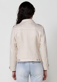 Jachetă de piele ecologică de culoare bej deschis, cu epauleți pe umeri, design strâns și detalii metalice argintii, asociată cu blugi de denim de culoare albastru deschis.