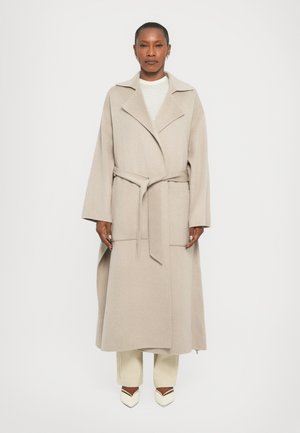 Beige long wool coat with wide sleeves, tie belt at waist, lapel collar, and front pockets. Worn over light-colored trousers and shoes.