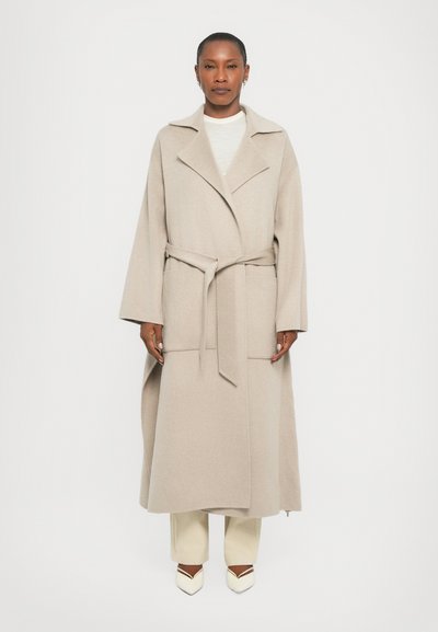 Manteau long beige en laine avec des manches larges, ceinture à nouer à la taille, col à revers et poches avant. Porté sur un pantalon et des chaussures de couleur claire.
