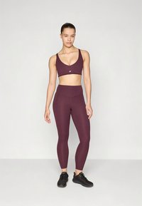 Športni modrček v rdečkasto rjavi barvi in visokopasne pajkice iz raztegljive tkanine. Prilegajoč dizajn z gladko teksturo in minimalnim blagovnim znakom na modrčku.