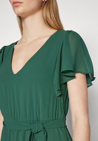 Rochie verde cu decolteu în V, mâneci fluturând și talie legată. Țesătura pare ușoară și netedă, cu o cădere subtilă.