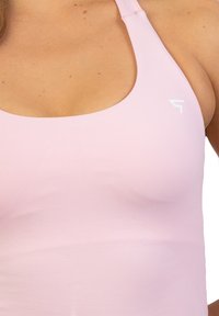Jasnoróżowy stanik sportowy z okrągłym dekoltem i wąskimi ramiączkami. Posiada subtelną teksturę oraz małe białe logo w prawym górnym rogu.