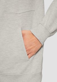 Detalle de sudadera gris que muestra una tela texturada, bolsillo lateral y manga con puño. El material parece suave e incluye acentos acanalados.