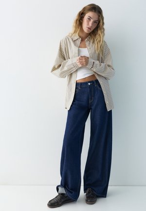 Giovane donna con lunghi capelli ondulati che indossa una camicia beige a quadri sopra un top corto bianco, jeans a gamba larga blu scuro e scarpe nere, in piedi.