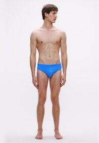 Μπλε μαγιό σλιπ κατασκευασμένα από ελαστικό υλικό με σχεδιασμό χαμηλής μέσης. Διαθέτουν λογότυπο στη ζώνη μέσης. Το μοντέλο στέκεται σε ουδέτερη στάση.