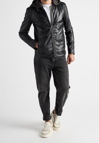 Černá kožená bunda se šedou kapucí, zapínáním na zip a dvěma předními kapsami, nošená přes šedé džínové kalhoty a bílé tenisky.