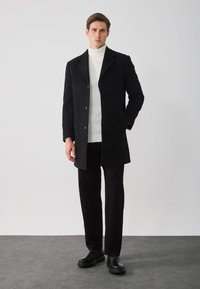 Abrigo de mezcla de lana negro con cuello de muesca, cierre de dos botones, llevado sobre un cuello de tortuga de punto blanco, combinado con pantalones y zapatos negros.