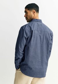 Koszula w granatowo-białą kratę z długimi rękawami, mankietami na guziki oraz klasycznym kołnierzem, z zakrzywionym dołem i detalem karczka z tyłu.