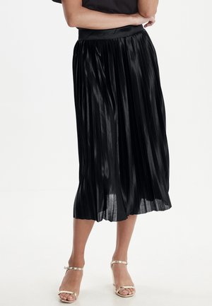 Czarna plisowana spódnica midi noszona z złotymi szpilkami na kostkę, pokazana na osobie z założonymi skrzyżowanymi rękami i czarnym topem.