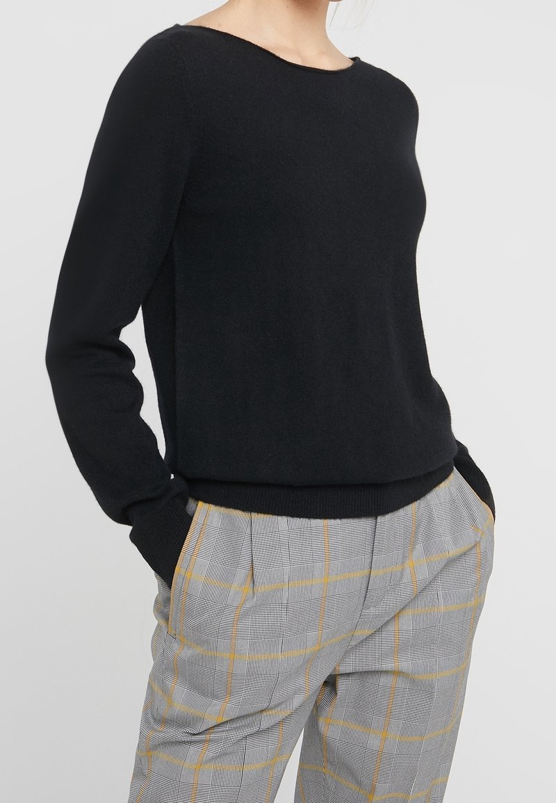 Pull en tricot noir avec un col rond et des manches longues. Associe à un pantalon à carreaux gris avec des accents jaunes et une coupe décontractée.