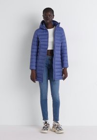 Casaco leve acolchoado em azul-marinho com capuz, apresentando um fecho de correr completo, mangas longas e um corte direito. Combinado com jeans de lavagem clara e ténis.