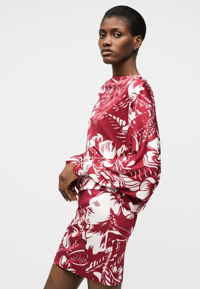 Γυναίκα με κοντά μαλλιά που φοράει μακρυμάνικο φόρεμα με κόκκινο και λευκό λουλουδένιο μοτίβο, στέκεται πλαγίως μπροστά σε ένα μονόχρωμο φόντο.