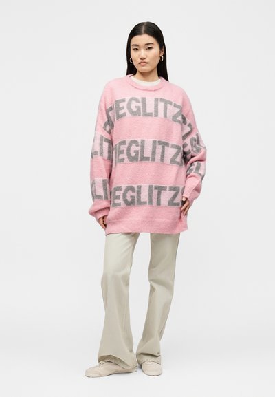 Femme debout portant un pull rose oversize avec le texte gris « GILLETZ », un pantalon beige large et des baskets beiges sur un fond uni.